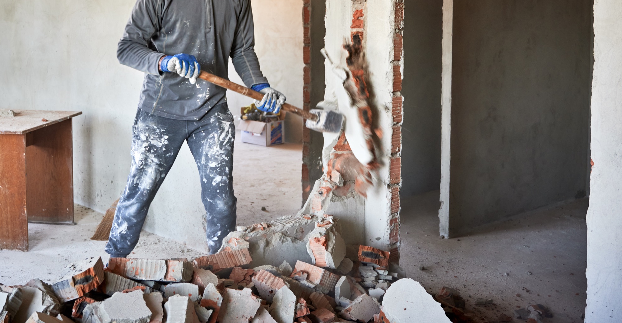 Worker swinging a sledgehammer through an interior brick wall during a demolition